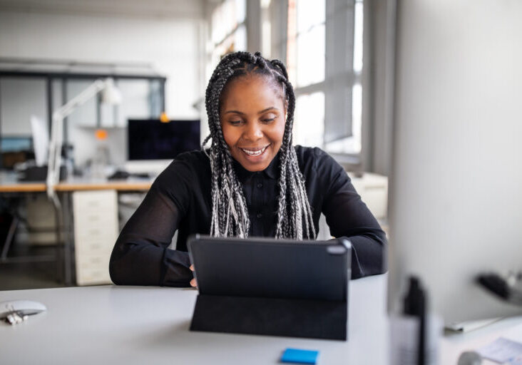 Smiling female executive sitting at her office desk making a video call with digital tablet. African american businesswoman working in office having a video conference.