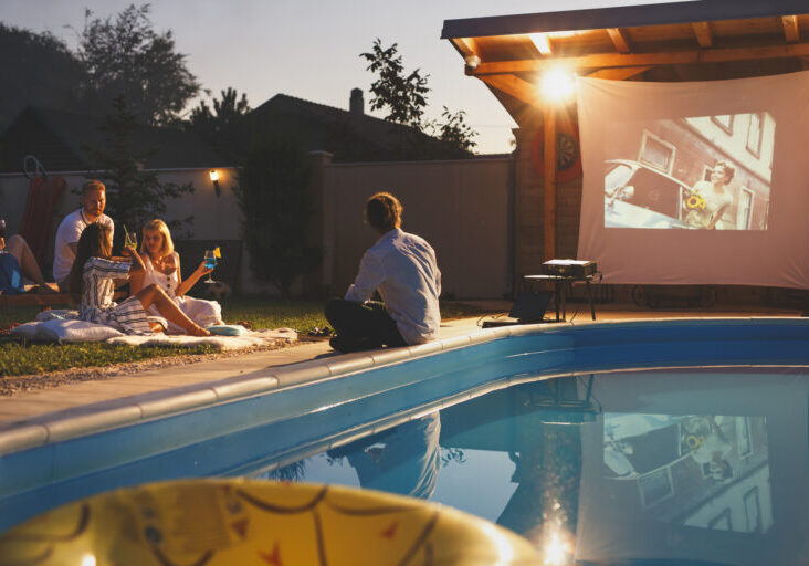 Group of cheerful young friends having fun eating popcorn, drinking cocktails and watching a movie in a home backyard poolside open air cinema
