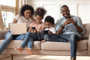 Happy young African American family with preschooler kids sit on couch use gadgets together